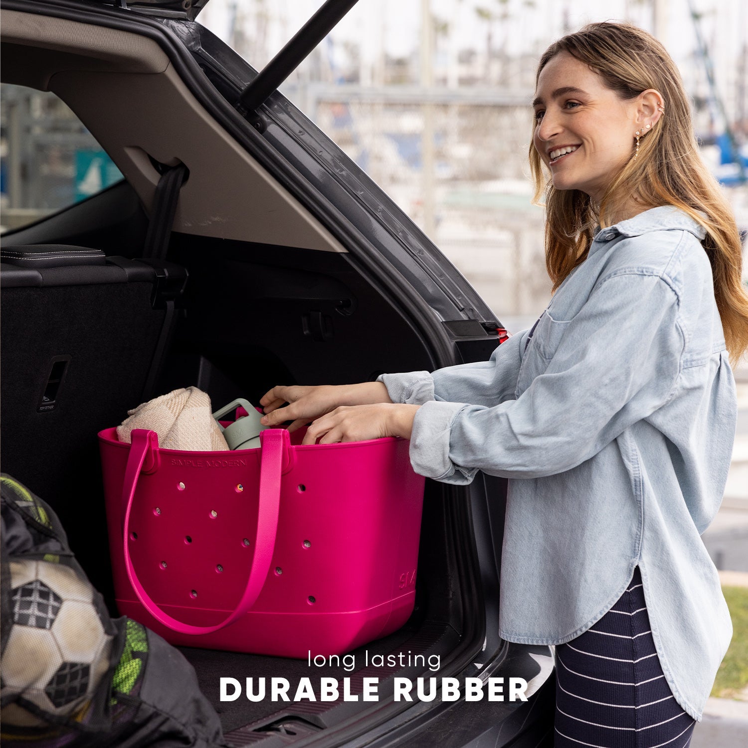 Woman placing a hot pink Simple Modern Getaway Bag into a car trunk, surrounded by towels and sports equipment. Text reads "long lasting DURABLE RUBBER." Urban background, daytime setting.