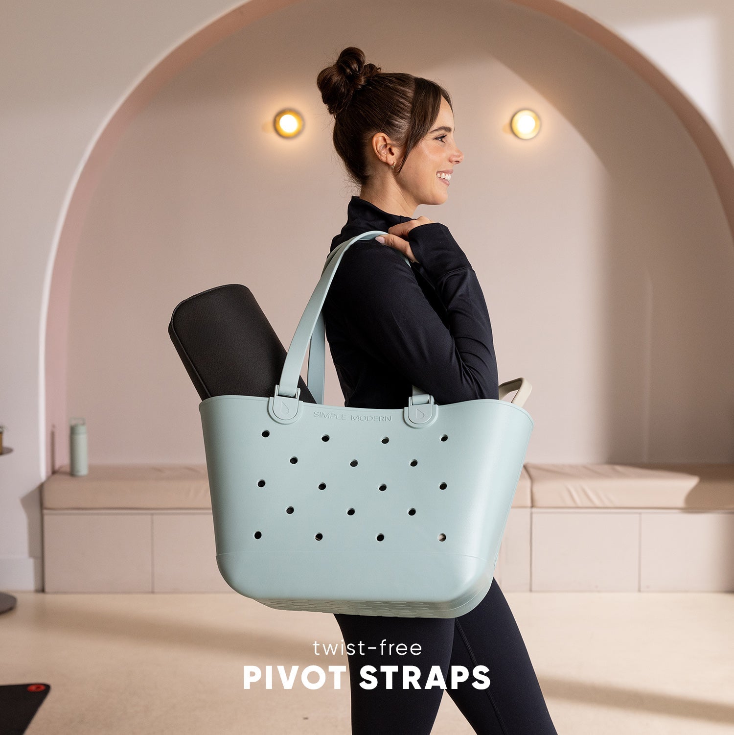 Woman wearing black top carrying light mint-green Simple Modern tote bag with perforated pattern and twist-free pivot straps in modern studio setting.