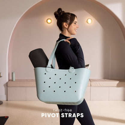 Woman wearing black athletic top and leggings carries a pale blue-green perforated tote bag with twist-free pivot straps over her shoulders in a modern studio setting with curved architecture.
