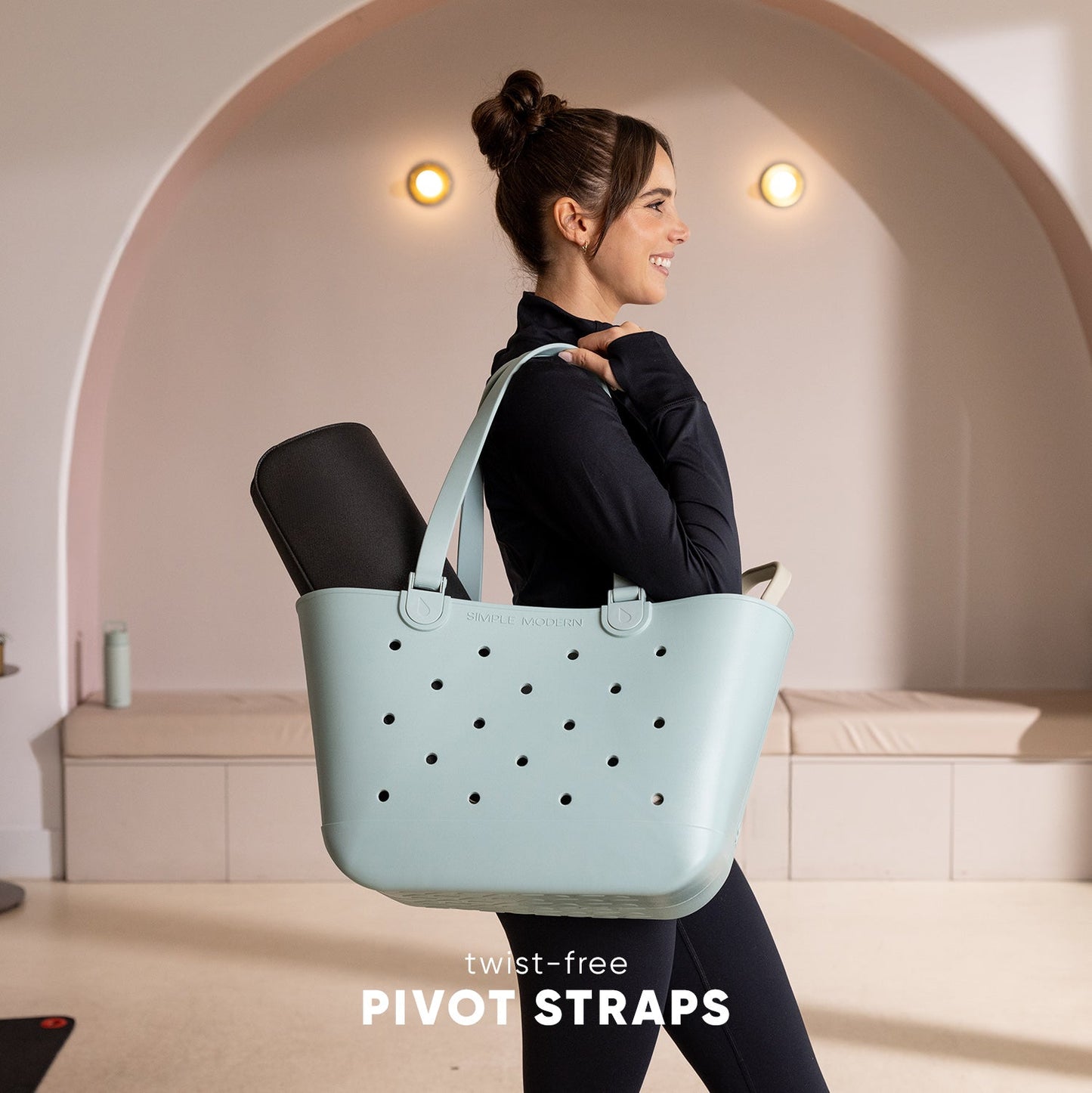 Woman in black athletic top carrying mint green Simple Modern Getaway Bag with perforated design and twist-free pivot straps in modern minimalist studio setting with arched alcove.