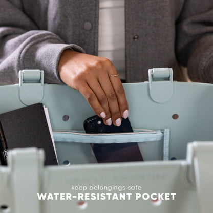Sage green tote bag with person placing black smartphone into water-resistant zippered pocket. Text reads "keep belongings safe" and "WATER-RESISTANT POCKET." Studio lifestyle product photo.