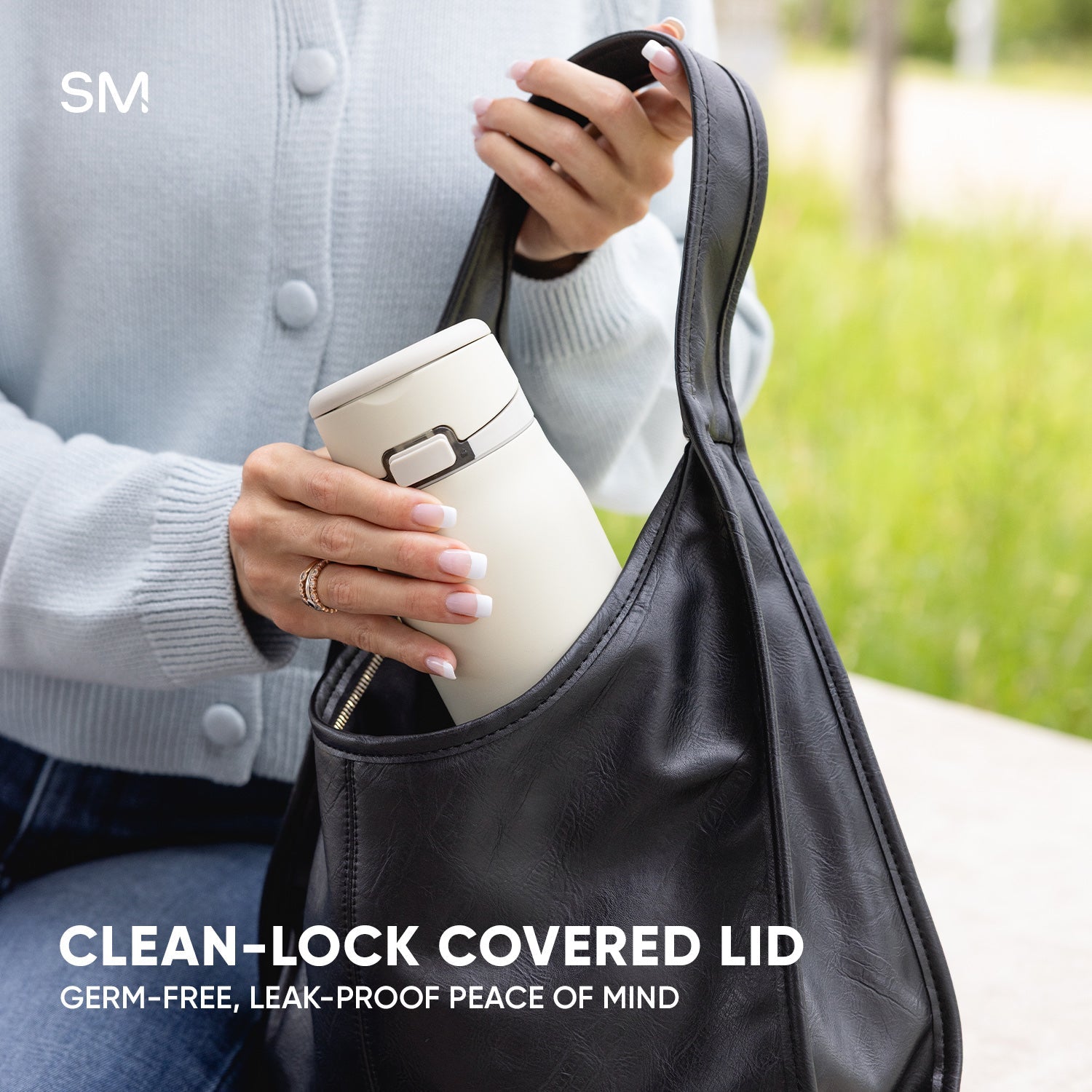 Person holding cream Simple Modern tumbler with Clean-Lock covered lid and metal buckle closure, wearing gray cardigan and carrying black shoulder bag outdoors in park setting.