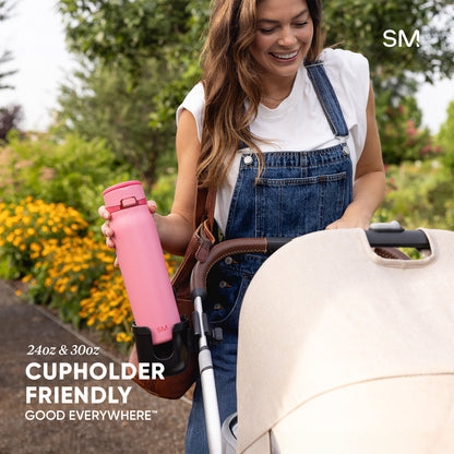 Woman in denim dress holding bright pink Simple Modern water bottle next to stroller in blooming garden. Text reads "24oz & 30oz Cupholder Friendly Good Everywhere."