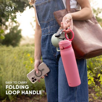 Person wearing blue denim vest holding pink Simple Modern water bottle with folding loop handle and carabiner, gray smartphone case, and brown leather bag in outdoor garden setting with green foliage.