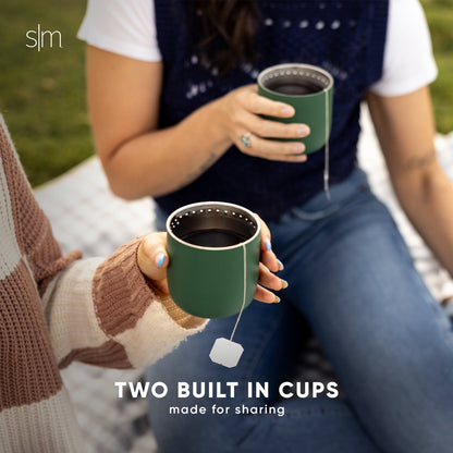 Two sage green insulated cups with white rim accents displayed alongside folded clothing in neutral earth tones and whites