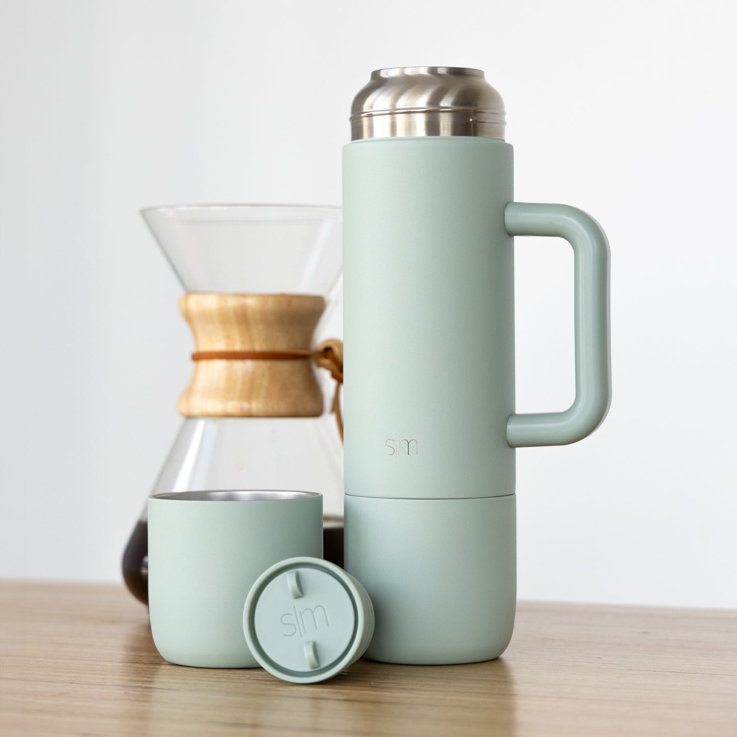 Sage green insulated thermos next to matching cup and Chemex coffee brewer with wood-tone band, on white background
